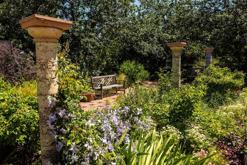 Stone columns by a wooden bench under leafy trees surrounding Denver Botanic Gardens at Warwick Denver