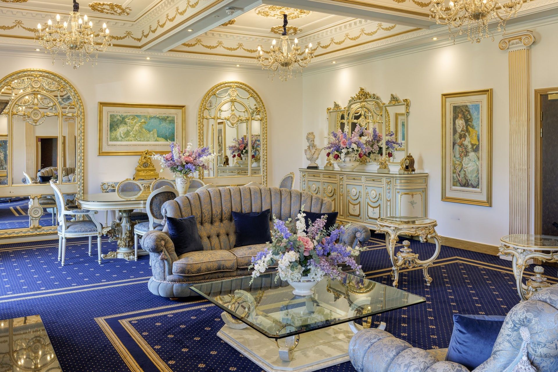 Ornate sofa and glass table between floral arrangements with grand gold mirrors and chandeliers at Polo Club Hotel