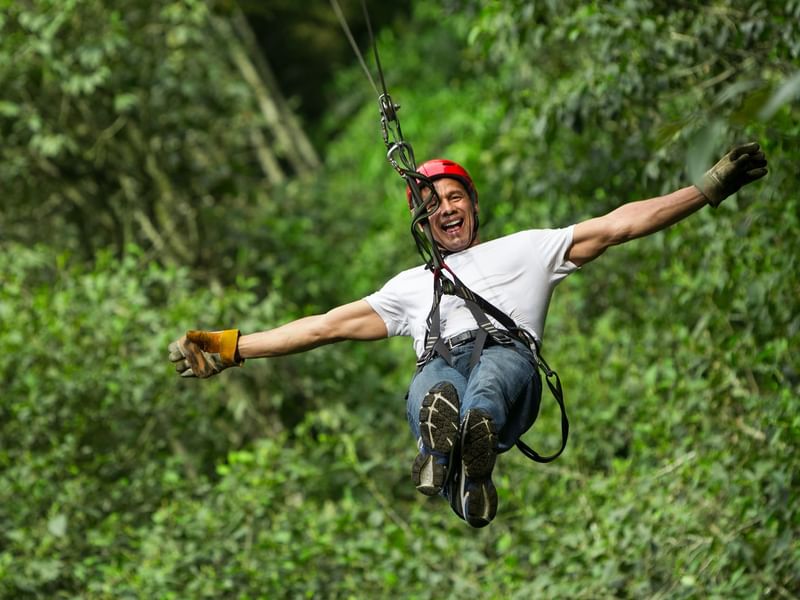 Person wearing red helmet and gloves zip-lining through lush green forest near Grand Fiesta Americana