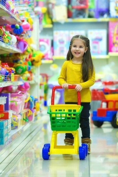 A little girl smiling while pushing a toy shopping cart down the aisle of a toy store.