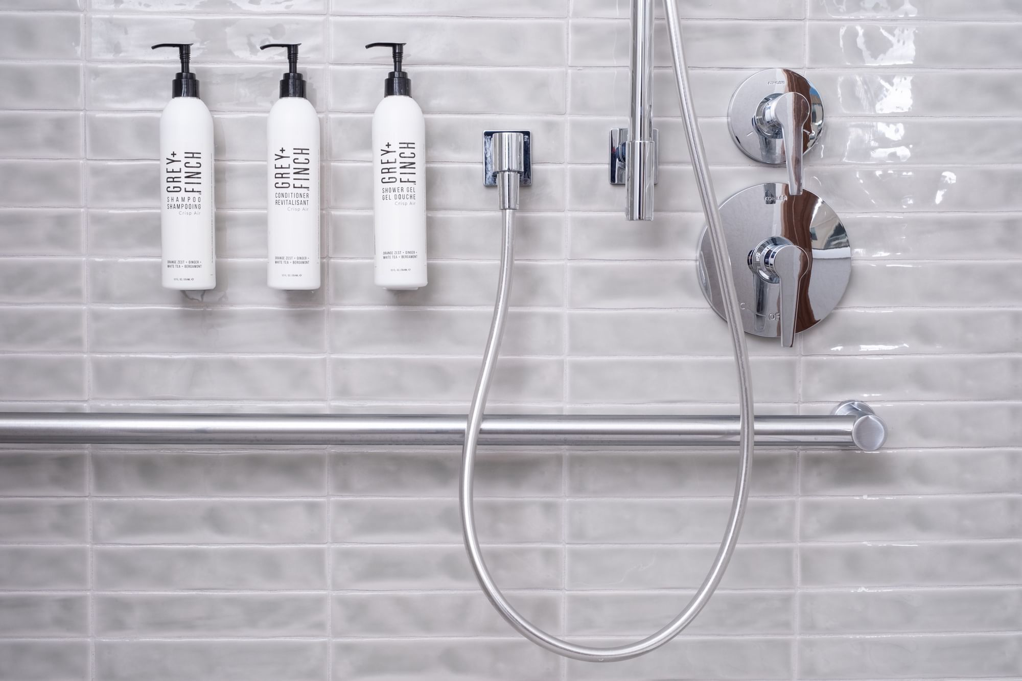 A shower with white tile walls, a silver shower head and controls, and three bottles of shower products.