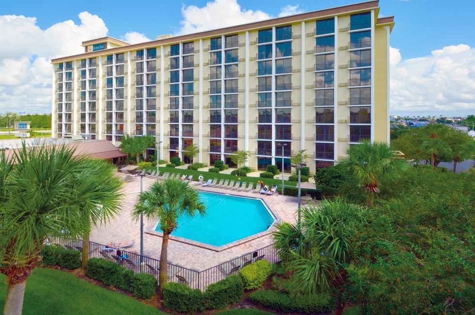 A tall hotel building against a blue sky stands beside a pool with a large patio, lounge chairs, and palm trees. 