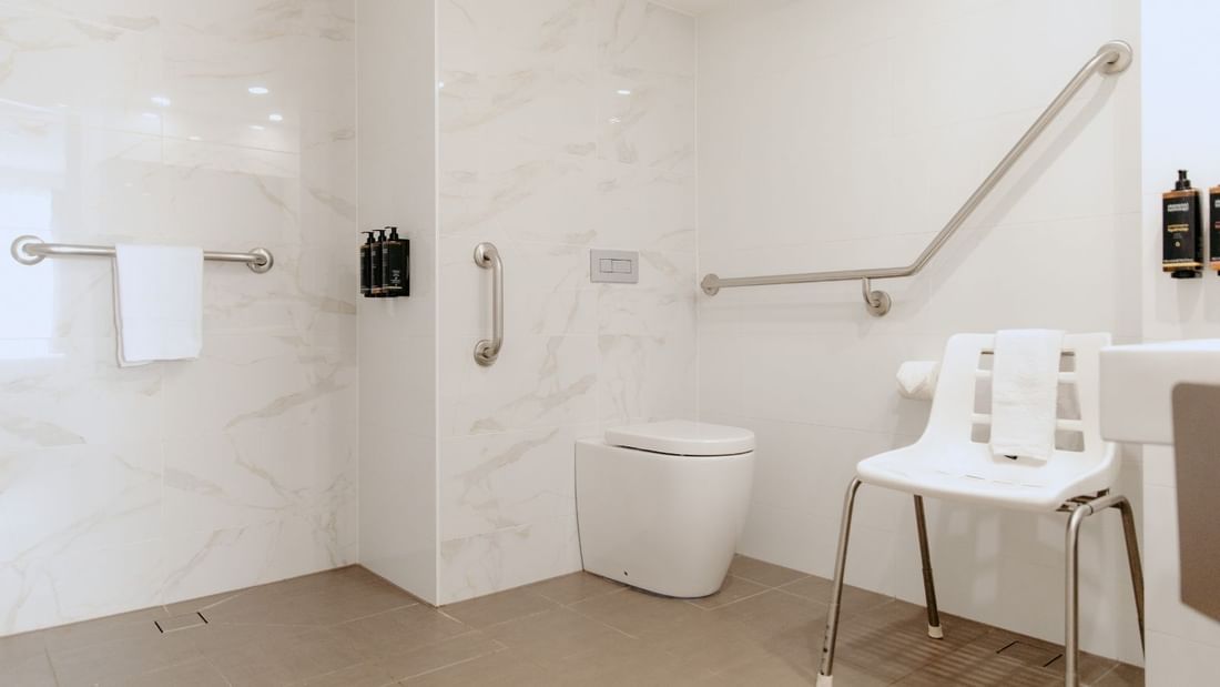 Accessible bathroom with white toilet, chair, and handrails, and a white towel hanging.