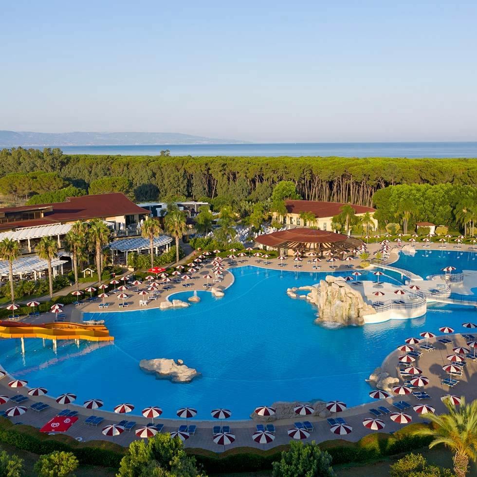 Luftaufnahme eines Resorts mit großen Pool, Liegen, Sonnenschirmen und umliegenden Gebäuden.