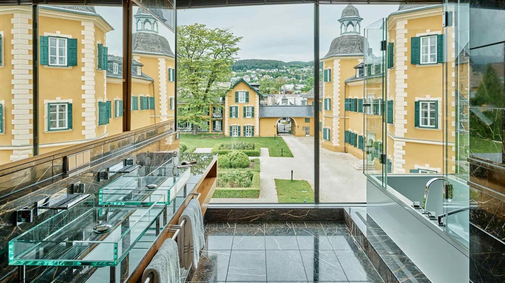 Glasbadezimmer mit Panoramablick auf gelbe Gebäude und gepflegten Garten.