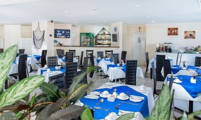 Modern restaurant with blue and white table settings, black chairs, plants, and a bar area at Bay Gardens Inn