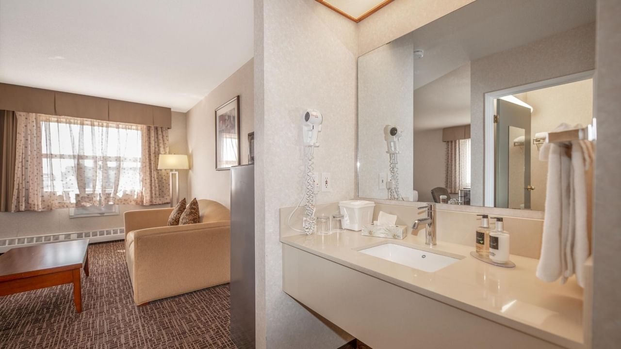 A hotel room with a couch, lamp, window, and a bathroom with a sink and mirror.