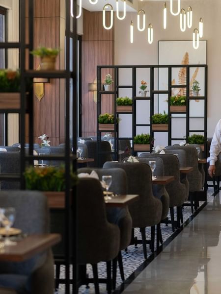Waiter carrying colorful juices through a dining area with gray chairs and metal shelving in La Maison at Warwick Al Jubail