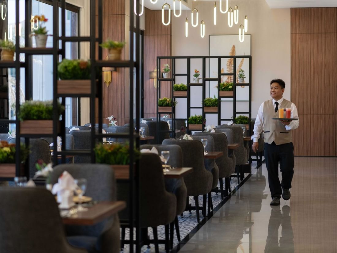 Waiter carrying colorful juices through a dining area with gray chairs and metal shelving in La Maison at Warwick Al Jubail