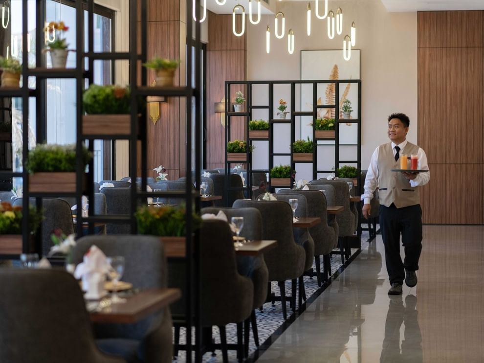 Waiter carrying colorful juices through a dining area with gray chairs and metal shelving in La Maison at Warwick Al Jubail