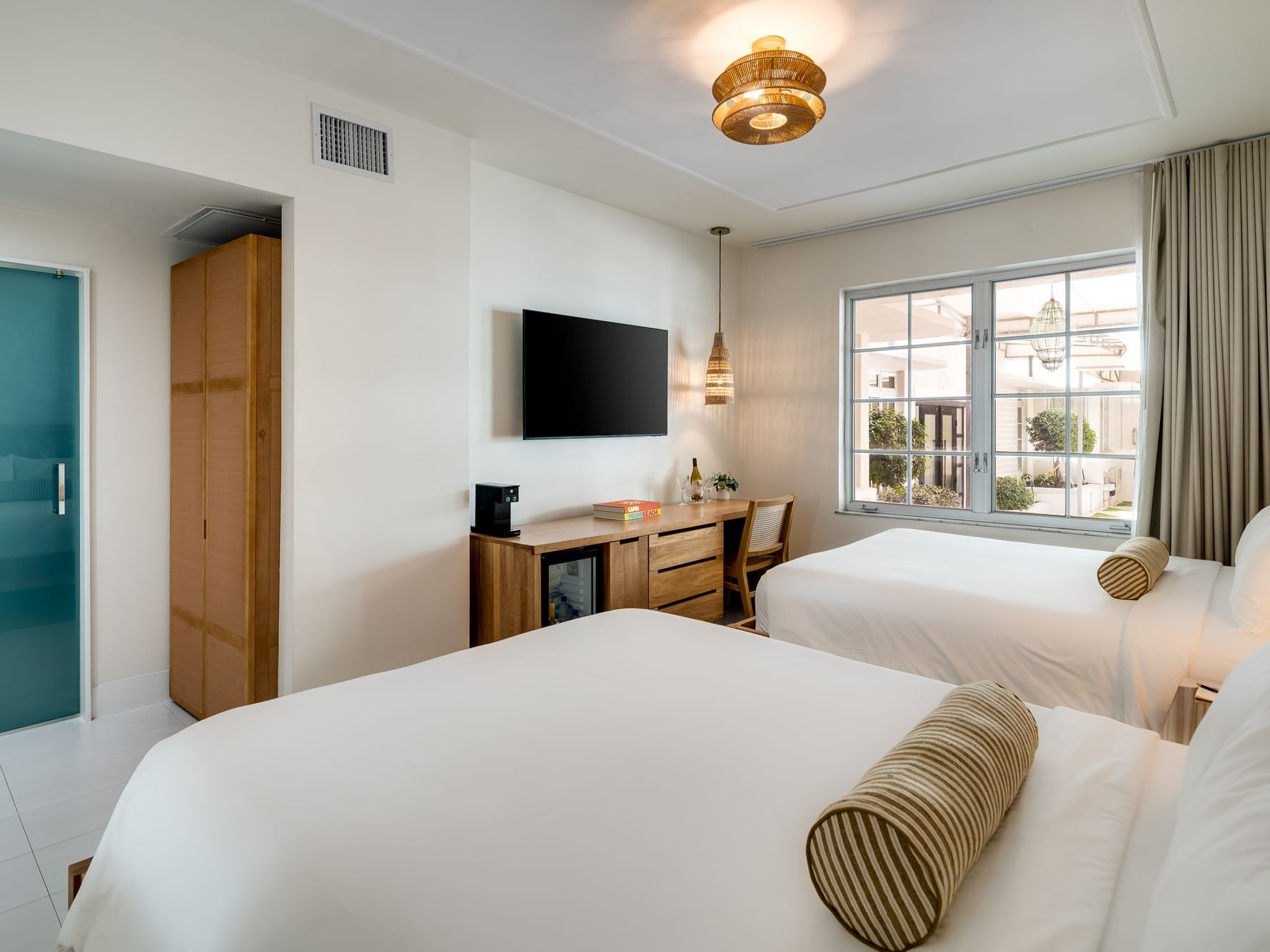 Newly renovated hotel room featuring two Queen beds with white linens and striped bolster pillows. The room includes a wall-mounted flat-screen TV, a wooden desk with a woven chair, a minibar with a coffee station, and pendant lighting that adds a touch o