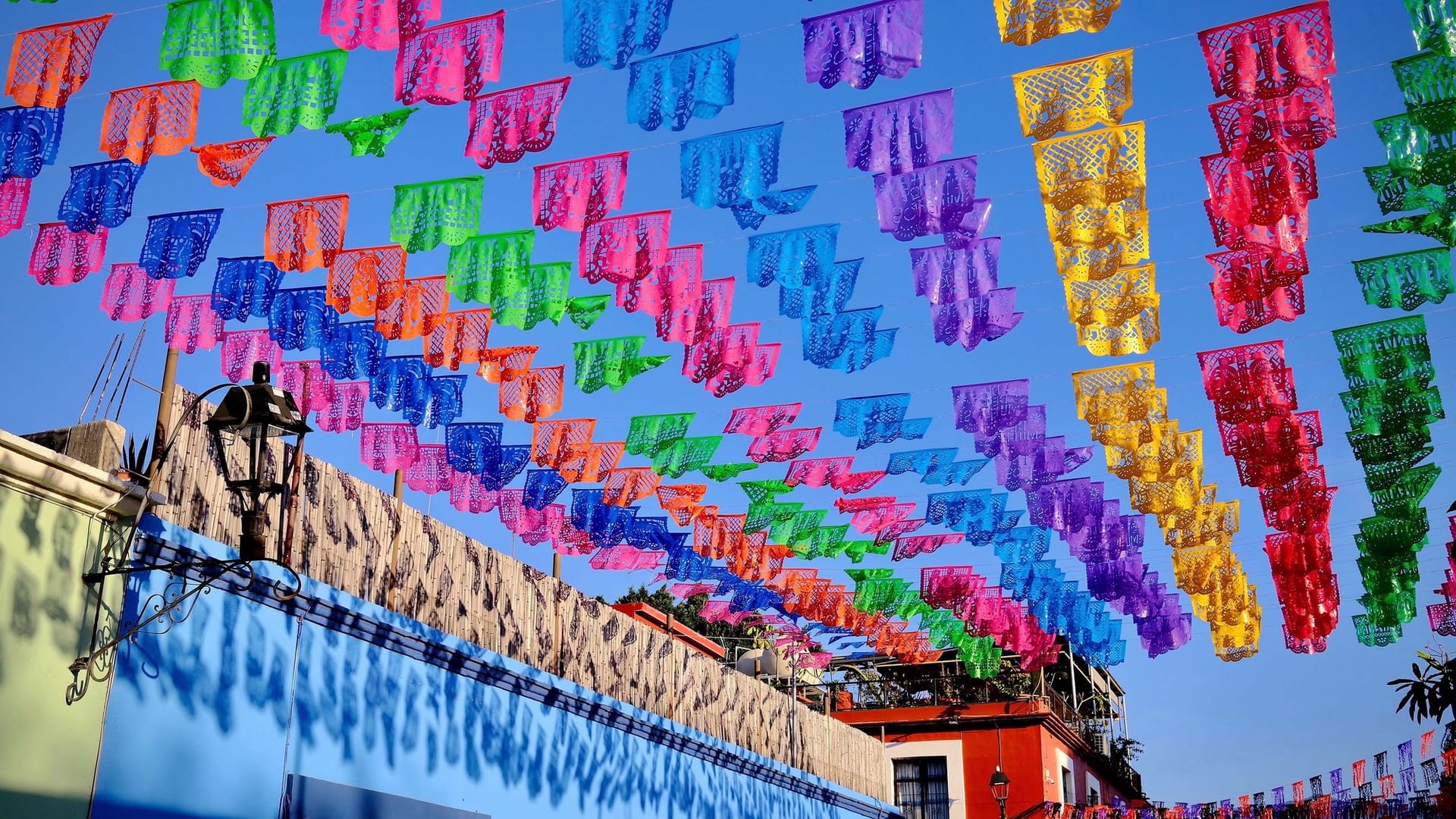 Coloridas banderas de papel picado colgadas sobre una calle, con cielo azul brillante y edificios debajo