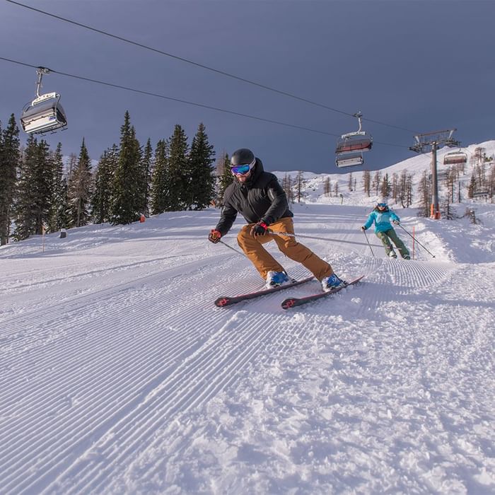 Zwei Skifahrer auf einer Piste mit Skiliften im Hintergrund, umgeben von Bäumen und Schnee.