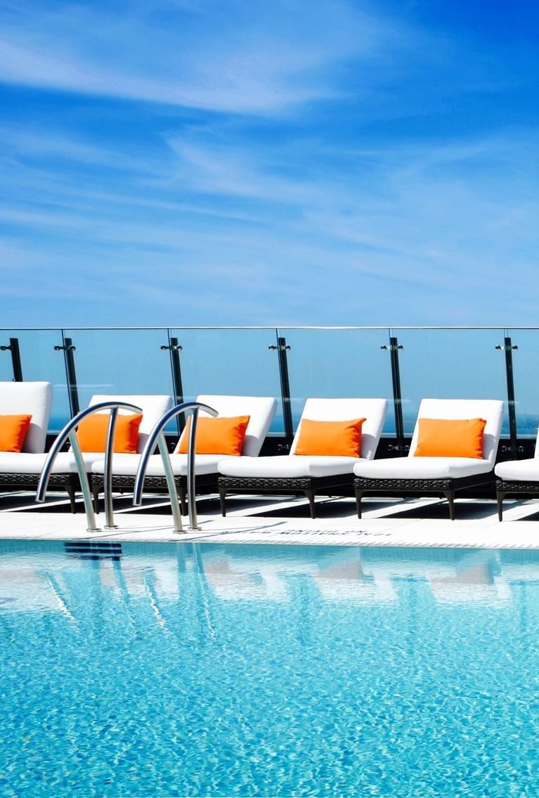 Row of white lounge chairs with orange pillows by a glass railing, overlooking a pool and skyline at Hotel X Toronto