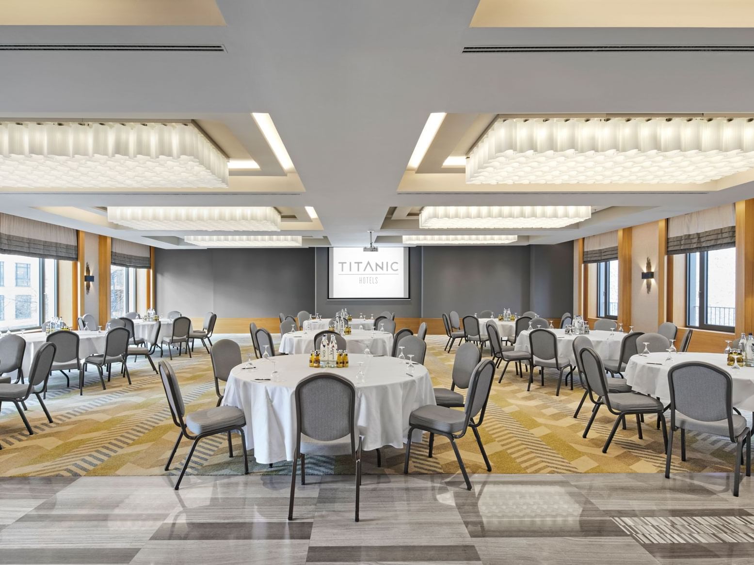 Konferenzraum mit mehreren Tischen, großen Fenstern und Wandbildschirm – Titanic Chaussee Berlin.