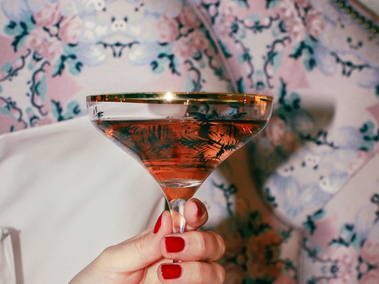 Hand with red nail polish holding a martini glass filled with a pink drink against a floral background.