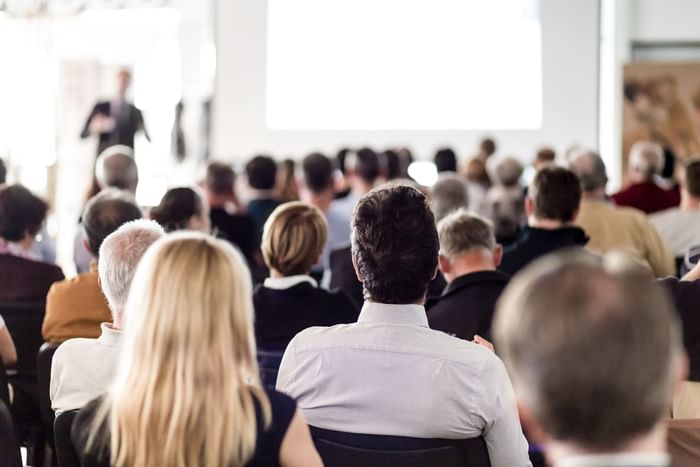 Audience members in a conference room with a speaker in front of a blank screen.