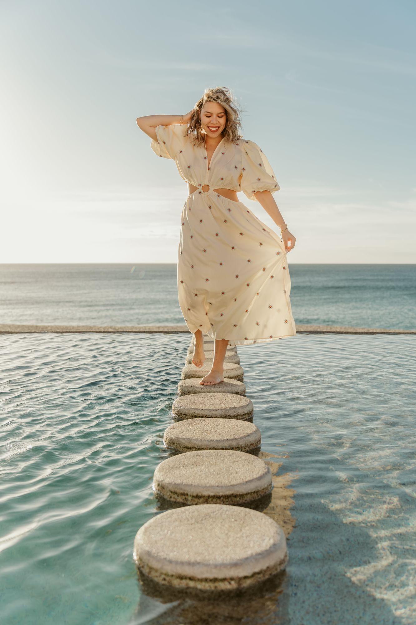 Woman in a flowing dress walking on stepping stones by the ocean at Marquis Los Cabos Resort