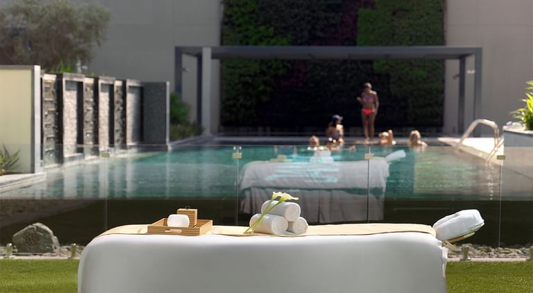 Luxury spa setting with massage table, towels, and serene pool with people relaxing at DAMAC Maison Cour Jardin