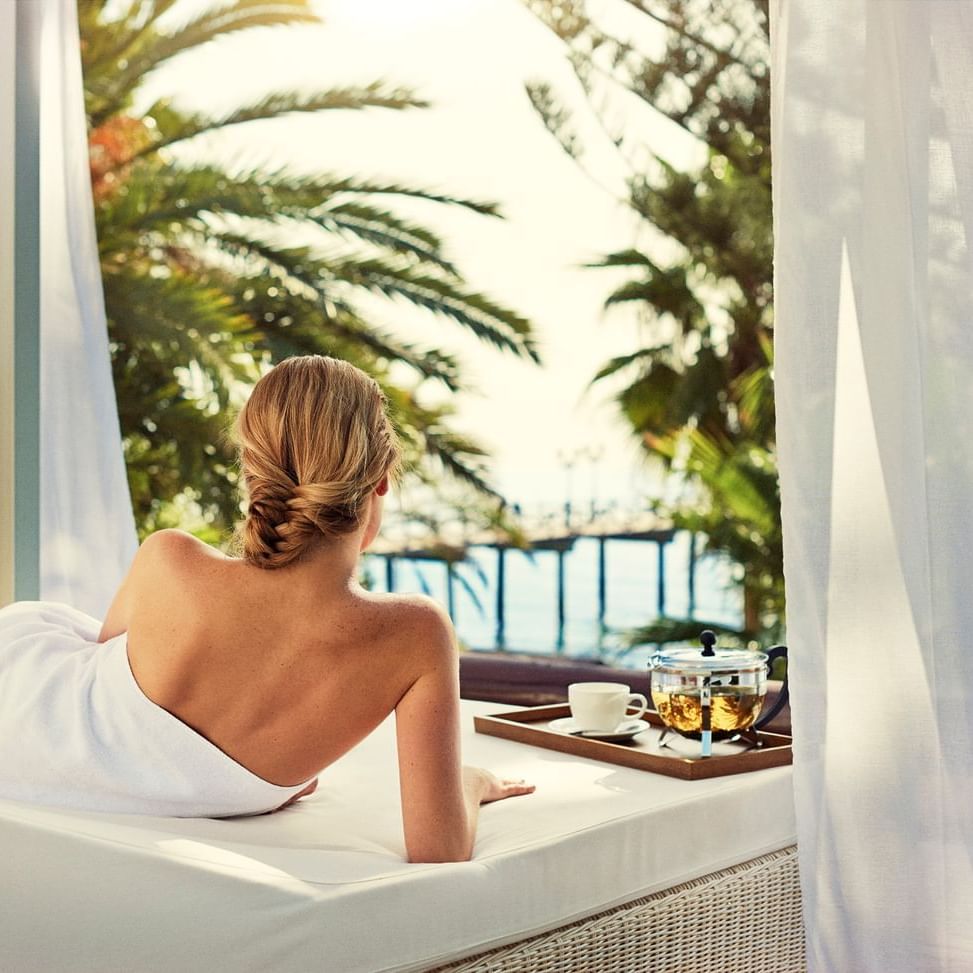 Woman in white towel relaxing on a bed with ocean view, surrounded by white curtains and palm trees.