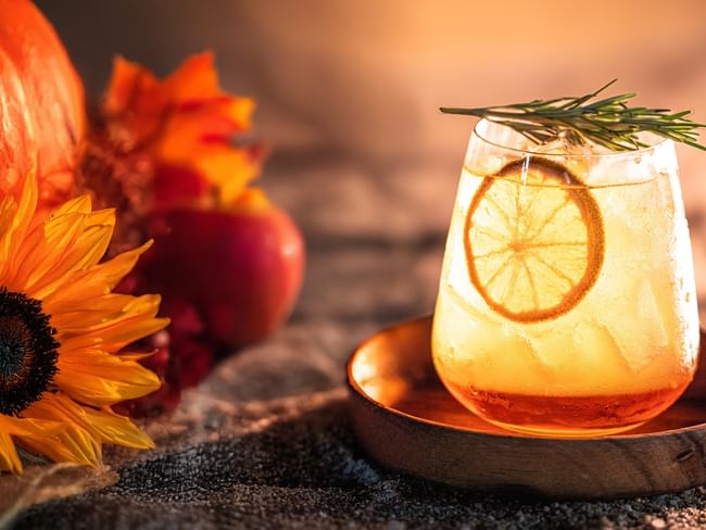 Glass of autumn drink with rosemary and lemon slice on a wooden plate, surrounded by sunflowers and a pumpkin.