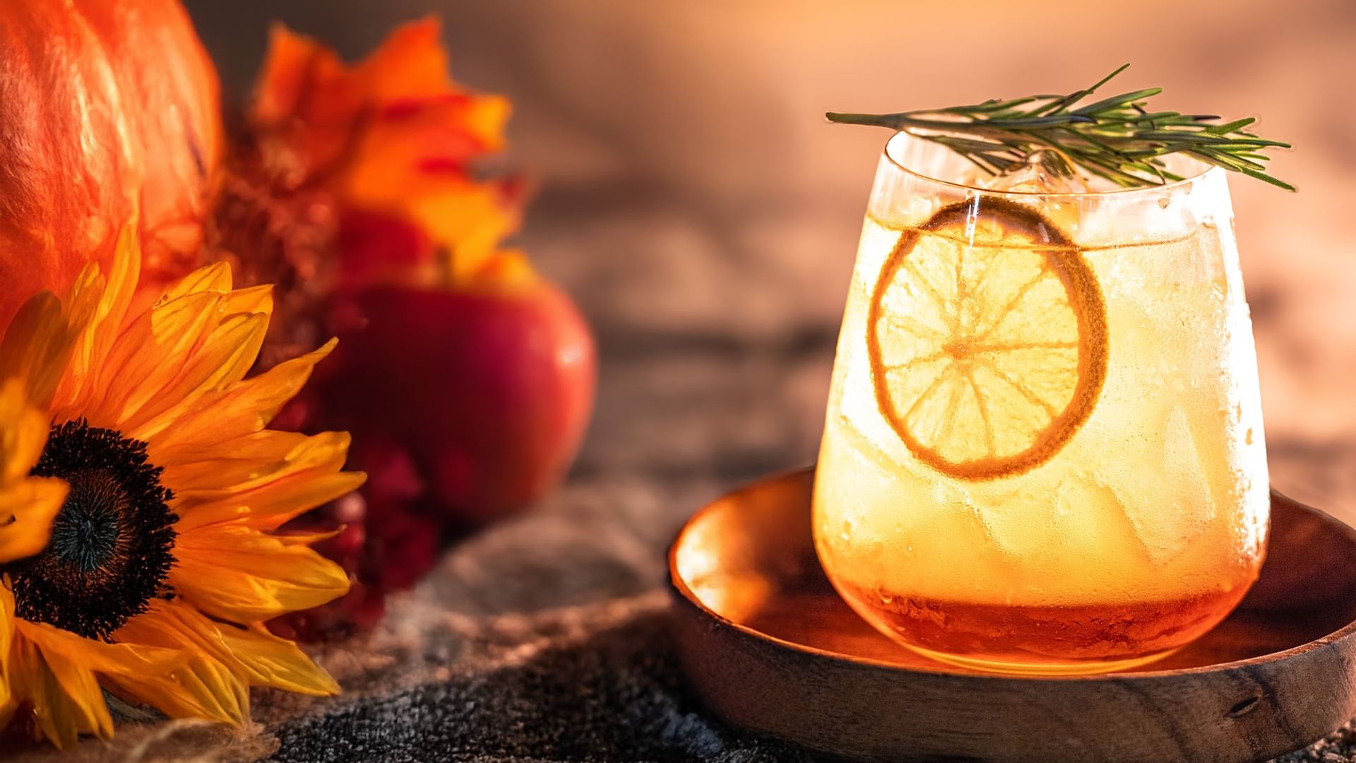 Vaso de bebida con rodaja de limón y romero, junto a calabaza y flores.