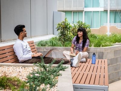 Two people sitting on benches in a landscaped terrace courtyard, with a building in the background.