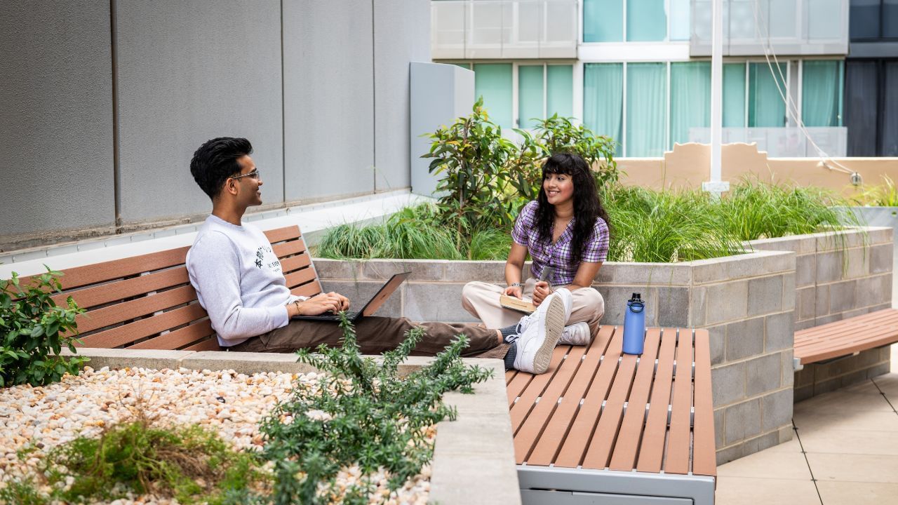 Two people sitting on benches in a landscaped terrace courtyard, with a building in the background.