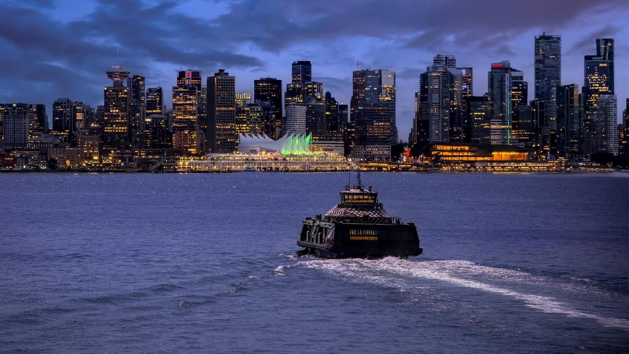 SeaBus ferry crossing harbor waters at dusk with illuminated city skyline in background.