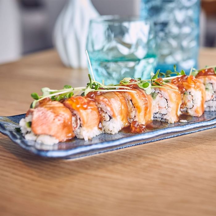 Sushi-Roller mit Reis, Lachs, Sauce und Kräutern auf einem Teller über einem Tisch mit Gläsern im Hintergrund.