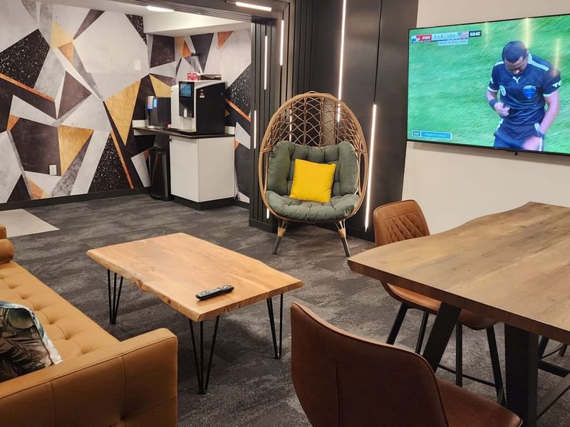 A lounge with sofa, coffee table, a worktable with stools, an egg chair, a 65-inch television, game boards and coffee station