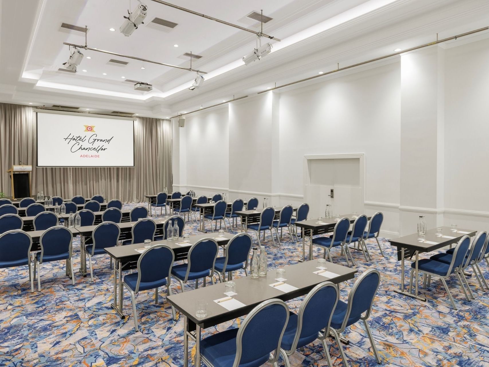 High-capacity conference room at Hotel Grand Chancellor Adelaide with classroom-style seating, stage, dual projection screens and professional lighting.