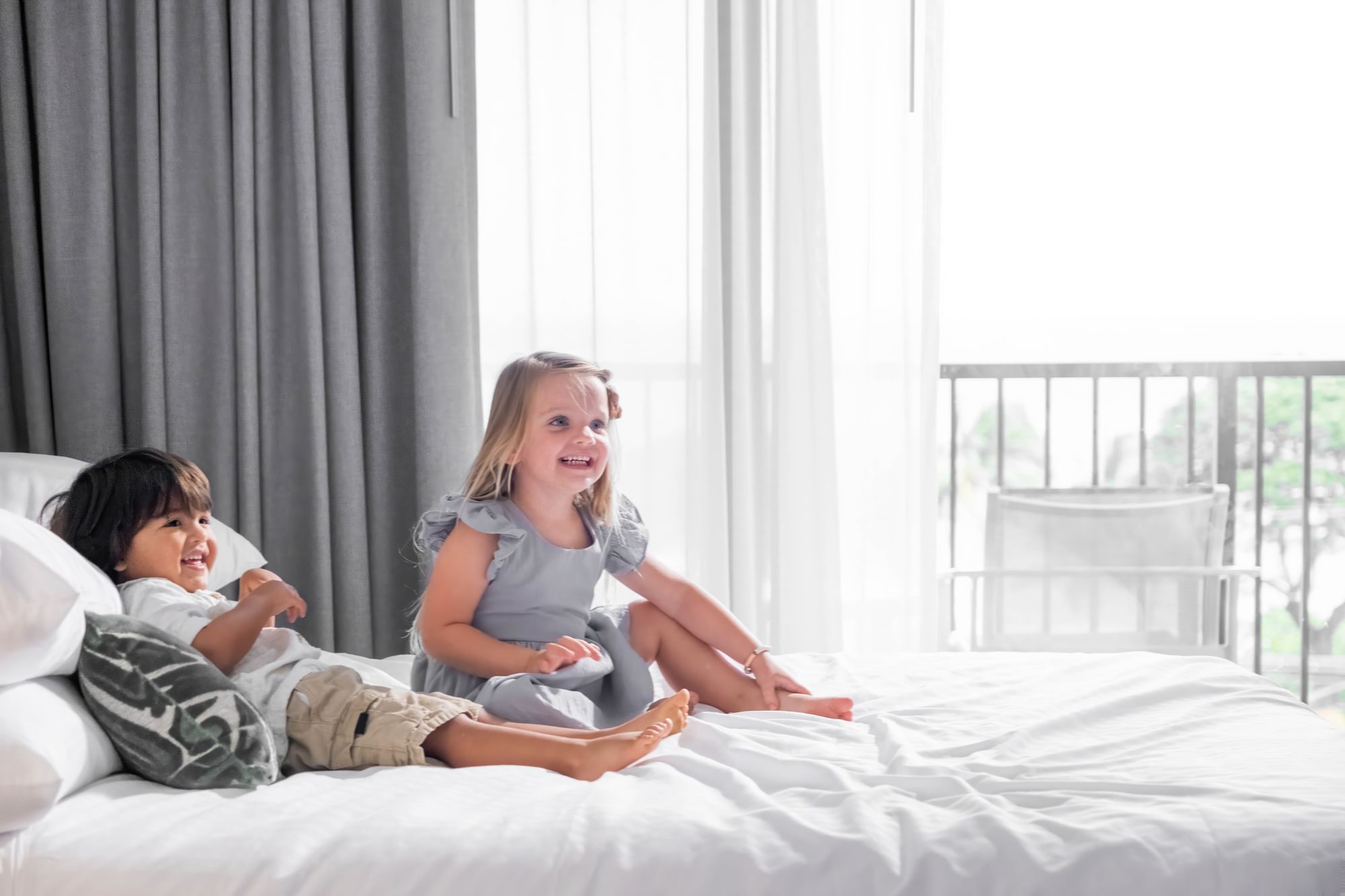 Two children sitting on a bed in a room with a large window and balcony.