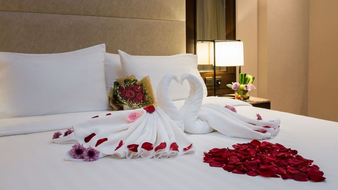 Towel swans with red rose petals on a white bed by a lamp in the Executive suite at Naviti by Warwick Dammam