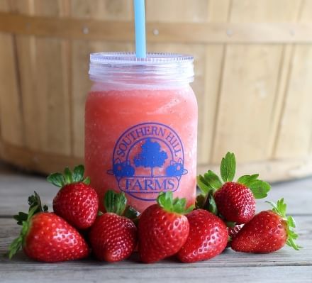 A mason jar with the blue Southern Hill Farms logo filled with a frozen pink drink sits behind a handful of ripe red strawberries. 