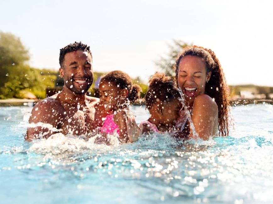 Family of four joyfully splashing in a pool, surrounded by greenery and a bright sky.