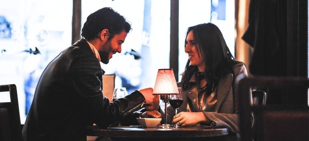 A couple sitting at a small restaurant table, smiling and talking over glasses of red wine in a warm, dimly lit setting.