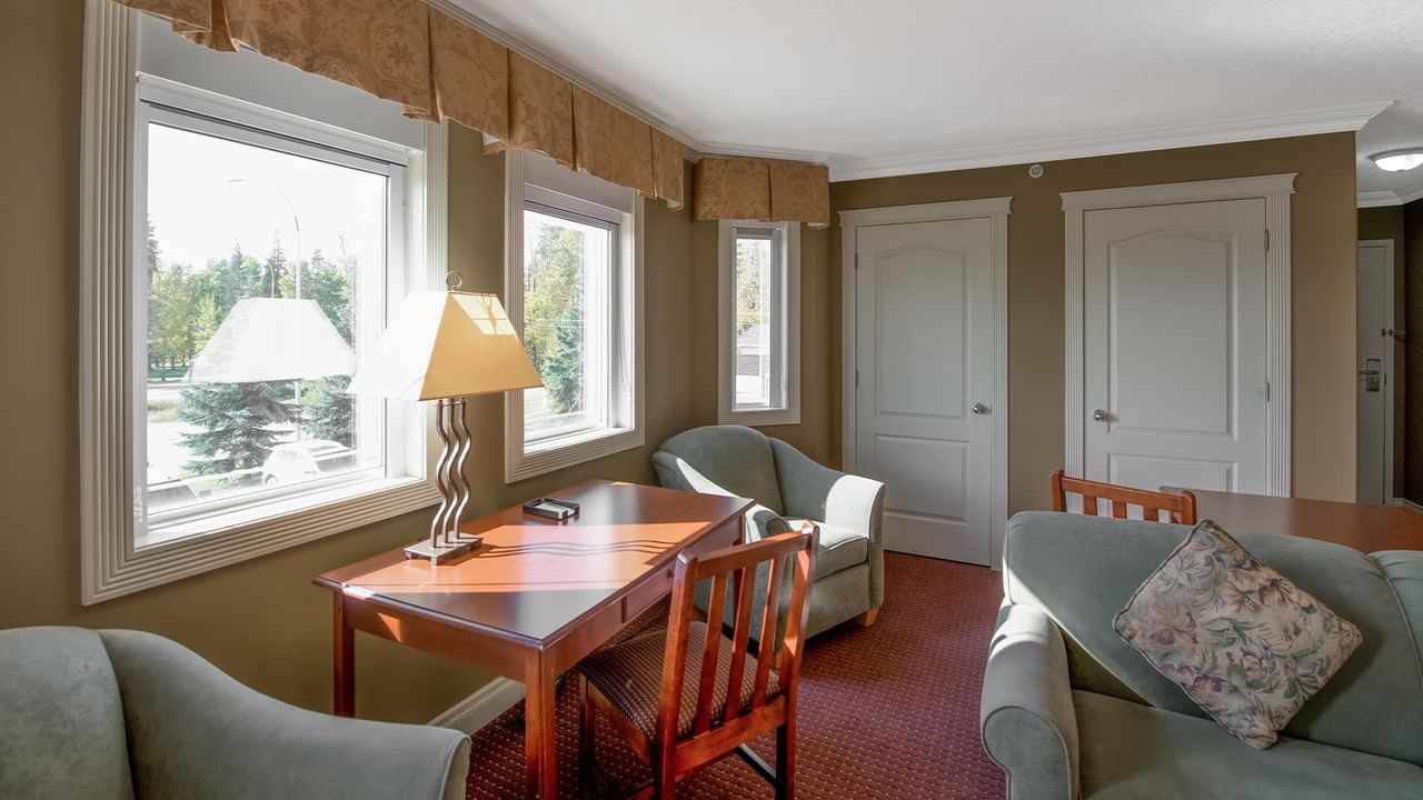 Coast Edson Hotel suite with wooden desk, teal armchairs, pendant lamps, and large windows overlooking greenery.