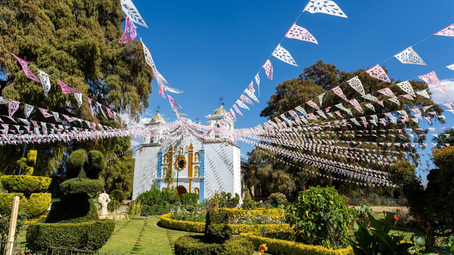 Iglesia blanca decorada con banderas rosas y jardín cuidado bajo el sol en Quinta Real Oaxaca