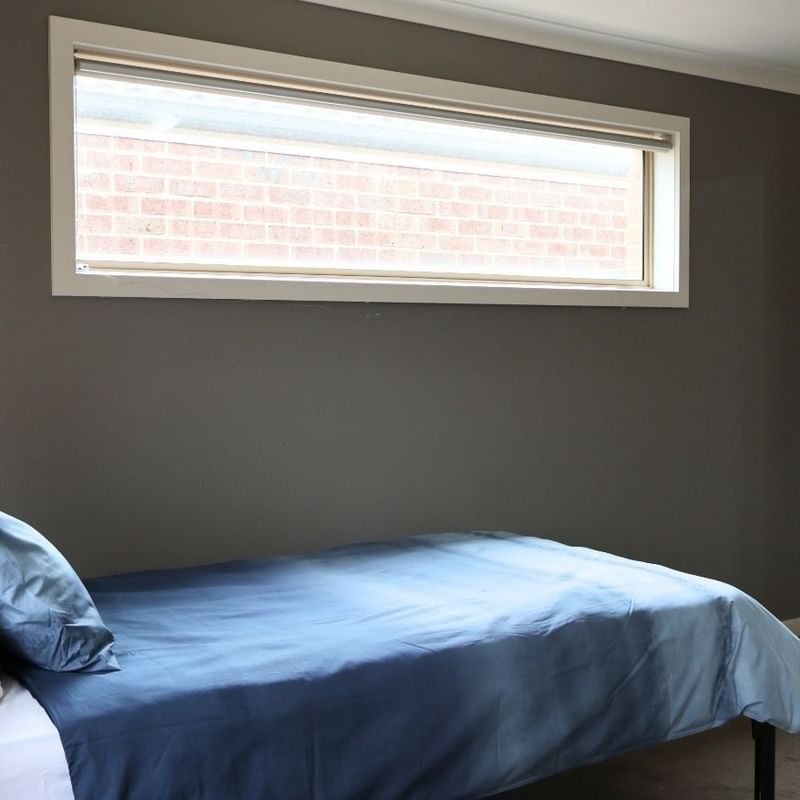 A cozy bedroom with a blue bed, gray walls, and a large window at La Trobe University Regional Housing – Melton.