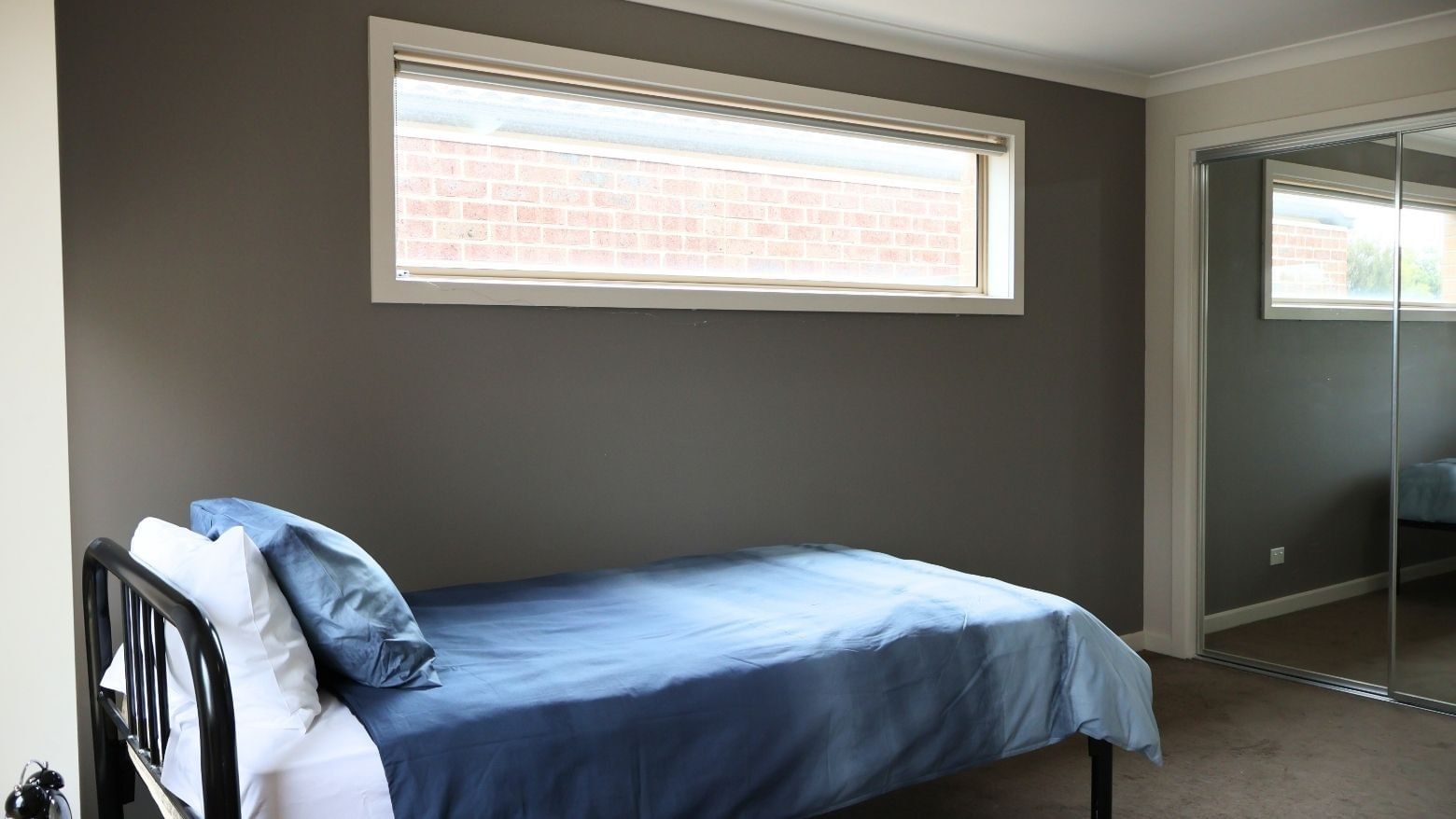 A cozy bedroom with a blue bed, gray walls, and a large window at La Trobe University Regional Housing – Melton.