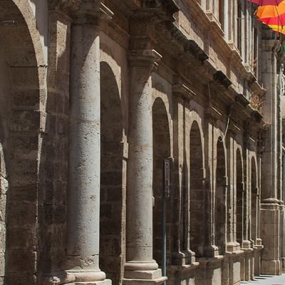 Bunte Regenschirme hängen über einer Straße vor einem Gebäude mit Säulen und Bögen.