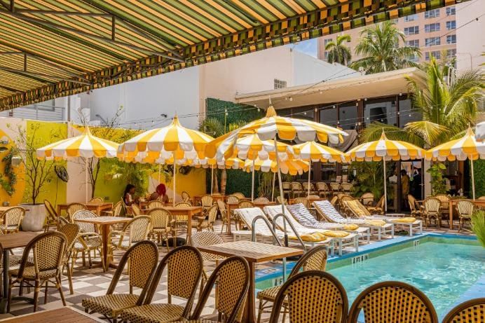 A poolside area with yellow and white striped umbrellas, brown wicker chairs, and wooden tables surrounding a swimming pool.