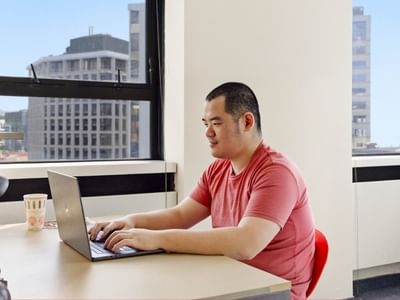 Man typing on a laptop at a desk with large windows in the background.