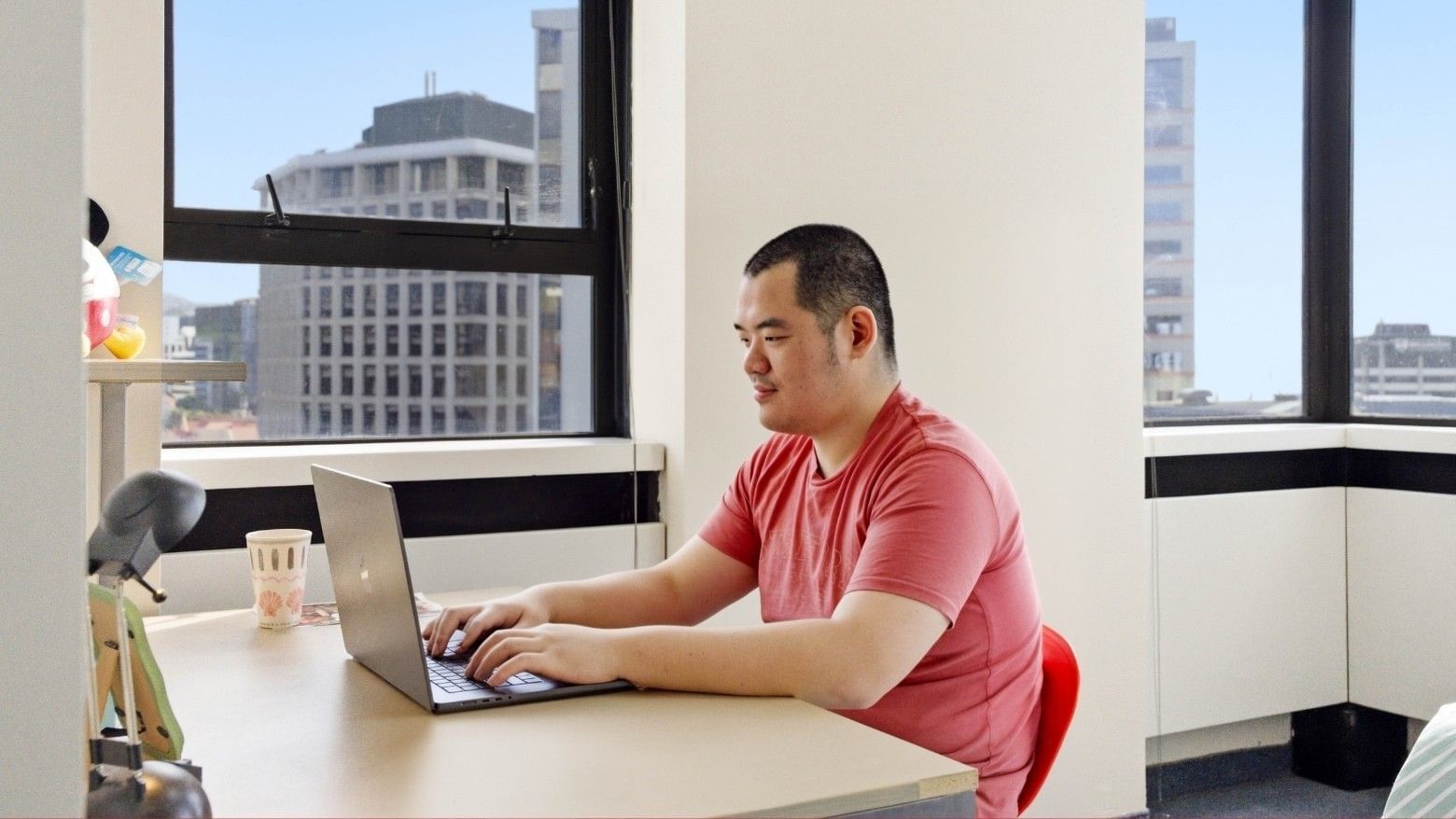 Man working on laptop at desk with large windows and city view.