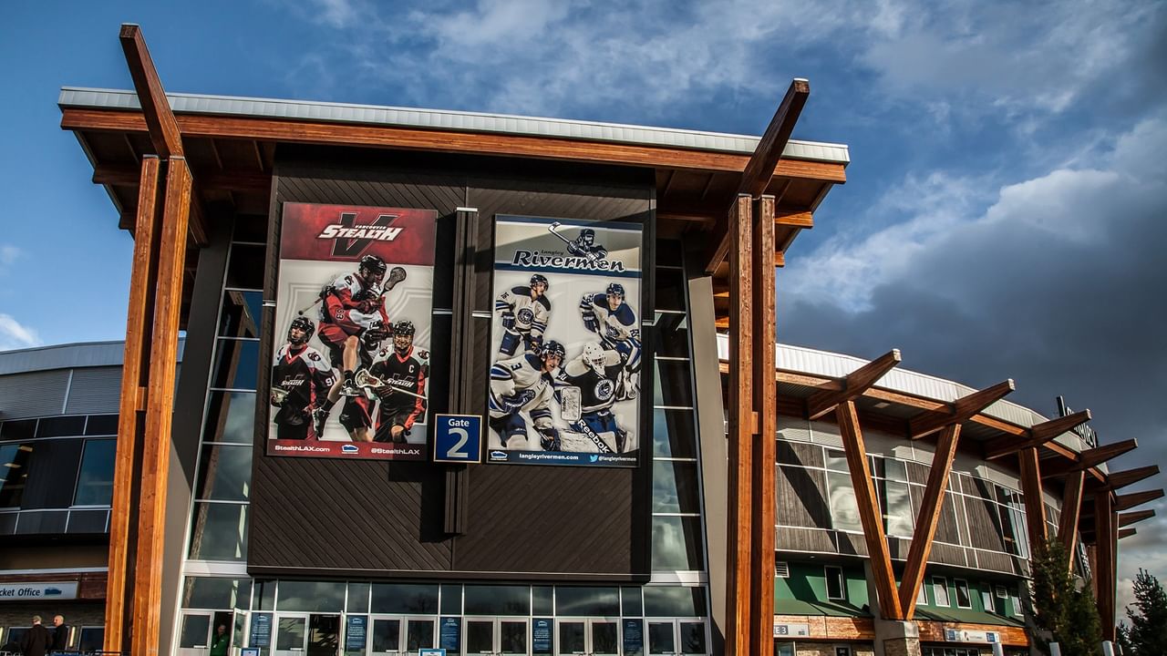 Exterior of sports arena with large banners of hockey teams, wooden supports, and cloudy sky.