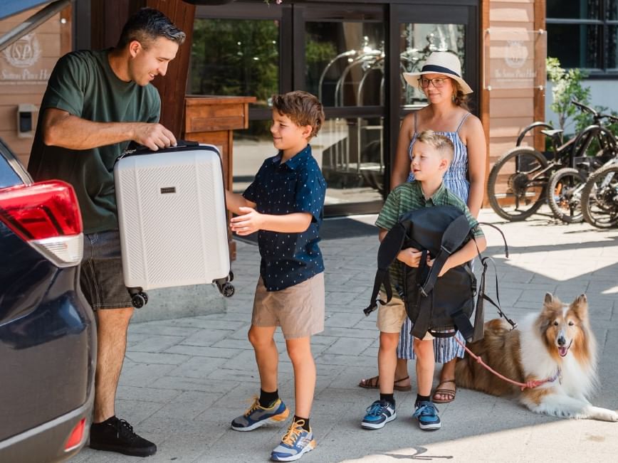 Family with kids and dog loading luggage into car for summer travel.