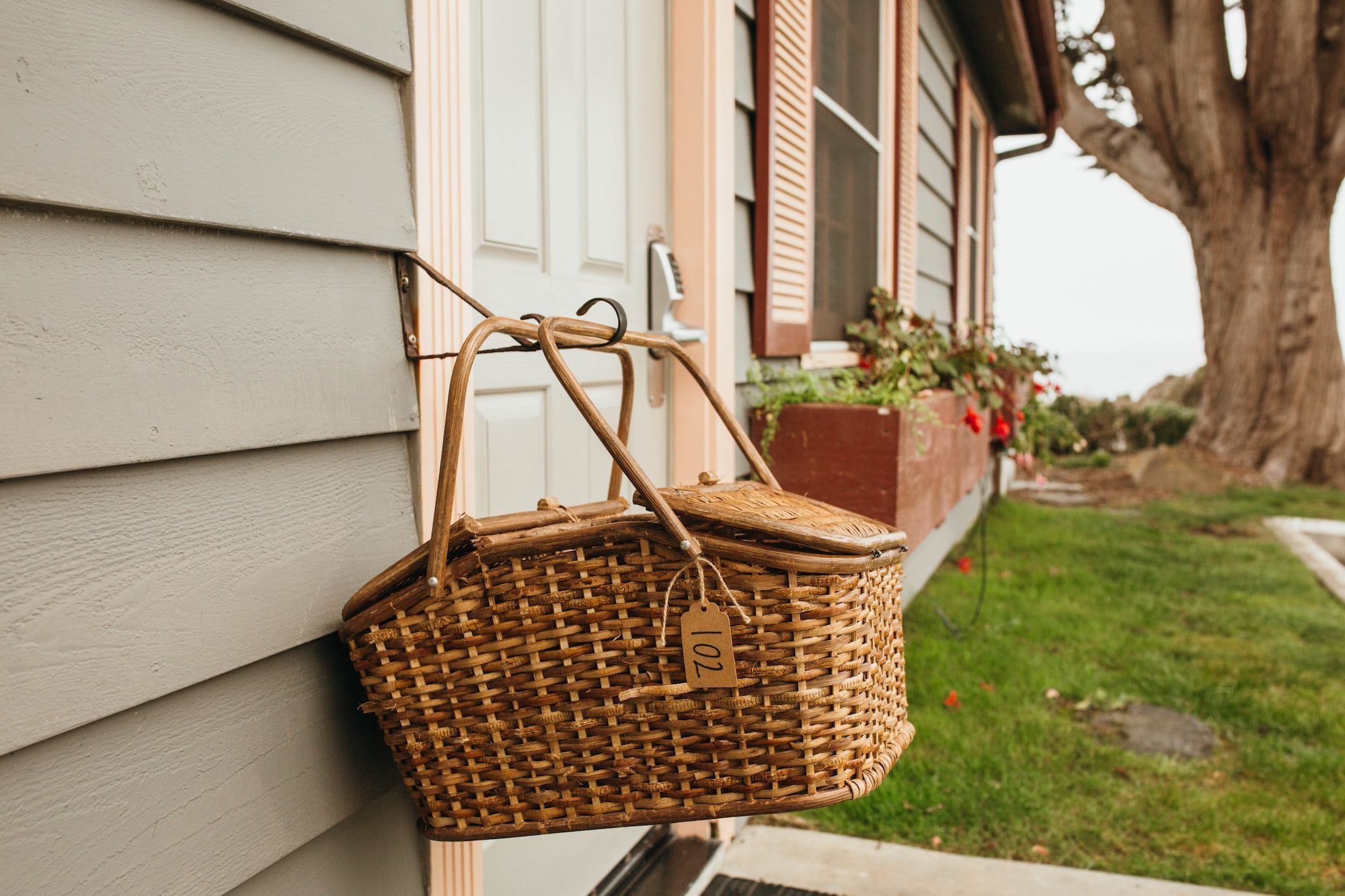 A wicker picnic basket hangs from a door handle on a house with gray siding, with a grassy area and tree in the background.