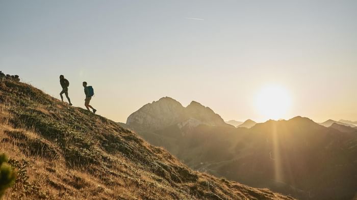 Due escursionisti con zaini camminano su una collina con montagne sullo sfondo al tramonto.