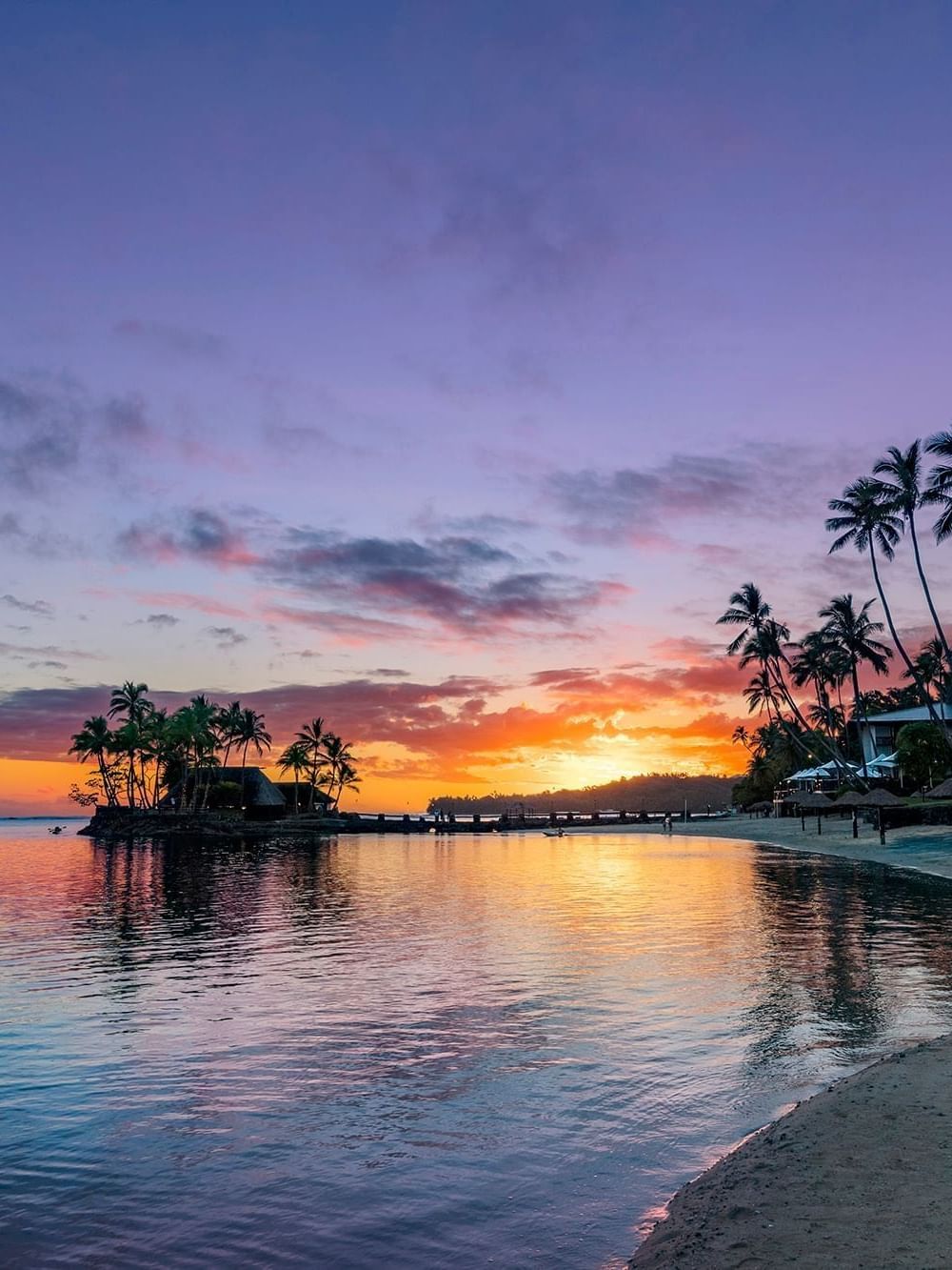 Des palmiers se découpent sur un coucher de soleil aux teintes violettes et orangées qui se reflète sur les eaux calmes du TokaToka Resort à Nadi, aux Fidji